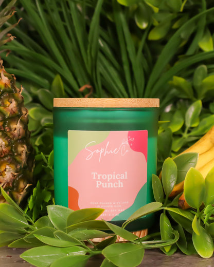 Hand poured soy wqax candle labeled 'Tropical Punch' by Sophie's Co. surrounded by greenery and a pineapple.